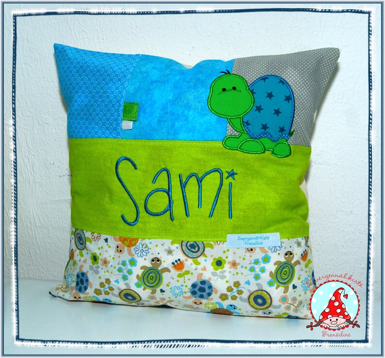 May include: A green and blue embroidered turtle is on a green and blue patchwork pillow with the name Sami embroidered on it. The pillow has a white background with a small turtle pattern.