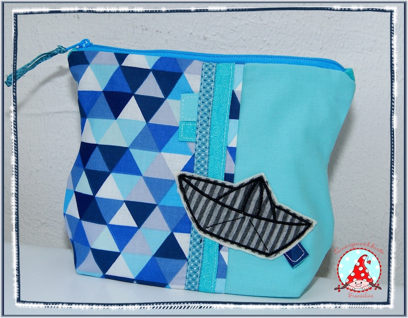 May include: A blue and white geometric pattern zippered pouch with a light blue body. A fabric applique of a paper boat is on the front. The pouch has a blue zipper and a blue fabric pull.
