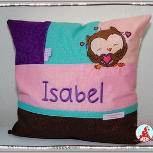 Könnte beinhalten: Ein rosa-braunes Kissen mit einem violetten und türkisfarbenen Patchwork-Design. Das Kissen ist mit einem gestickten Eulen, die ein Herz hält, und dem Namen "Isabel" verziert.