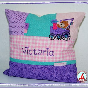 Könnte beinhalten: Ein lila, pink und türkisfarbenes Patchwork-Kissen mit einem darauf gestickten Zug. Der Zug hat einen braunen Bären vorne und der Name "Victoria" ist darunter gestickt.