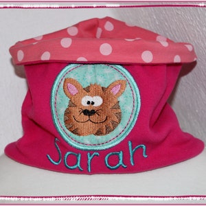 Könnte beinhalten: Ein rosa Fleece-Halswärmer mit weißem Punktmuster. Ein türkisfarbener Kreis mit einem braunen Comic-Katzenkopf und dem Namen "Sarah" in türkisfarbener Stickerei.