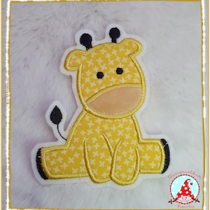 Könnte beinhalten: Ein gelber Giraffen-Aufnäher mit weißem Kreuzstichmuster, beiger Schnauze, schwarzen Augen, schwarzen Hornzapfen und einem schwarz gespitzten Schwanz. Die Stickapplikation hat einen weißen Filzrand und ist mit goldenem Garn umrandet.
