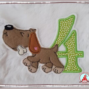 Könnte beinhalten: Ein brauner und hellbrauner Hundeaufnäher mit Knochen und der Zahl 4 in Grün mit weißen Punkten. Der Hund hat ein rosa Herz auf dem Ohr. Der Aufnäher befindet sich auf einem weißen, flauschigen Hintergrund.
