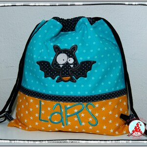 Könnte beinhalten: Eine türkisblaue Kordeltasche mit weißen Punkten. Die Tasche hat eine schwarze Fledermaus mit weißen Augen und Zähnen, die auf der Vorderseite gestickt ist. Der Boden der Tasche ist orange mit weißen Sternen und der Name "LARS" ist in blau gestickt.