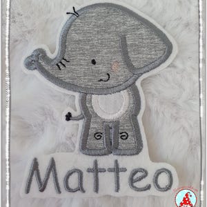 Könnte beinhalten: Ein gestickter grauer Elefant mit weißem Bauch und dem Namen Matteo darunter.