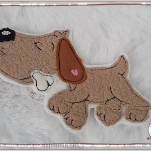 Könnte beinhalten: Eine braune Filzapplikation eines Hundes mit einem braunen Filzkragen und einem rosa Herz. Der Hund hält einen weißen Knochen im Maul.