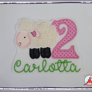 Könnte beinhalten: Ein weißes Schaf mit schwarzen Hufen und einer rosa Nase, eine rosa Zahl 2 mit weißen Punkten und der Name "Carlotta" in Grün auf weißem Hintergrund gestickt.