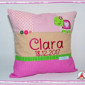 May include: A pink and beige pillow with a green and white striped border. The pillow has a green turtle with a pink heart on its shell and the name "Clara" embroidered in pink with the date "18.12.2017" below it.