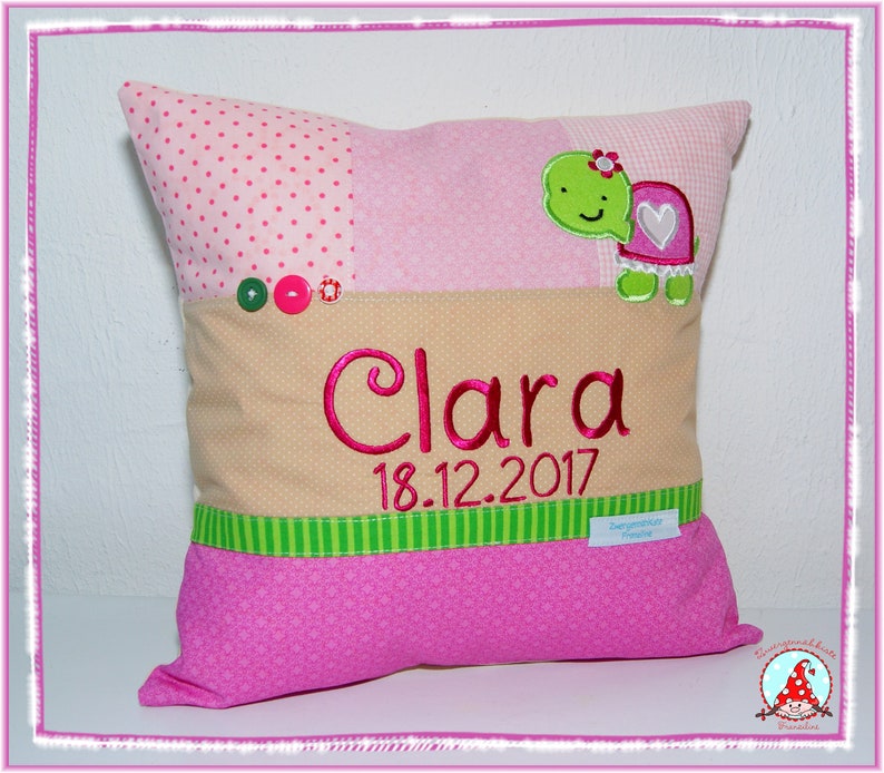 May include: A pink and beige pillow with a green and white striped border. The pillow has a green turtle with a pink heart on its shell and the name "Clara" embroidered in pink with the date "18.12.2017" below it.