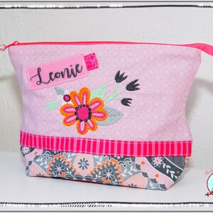 May include: Pink floral patterned zippered pouch with an embroidered flower design. The pouch has a pink zipper and a label that says "Leonic". The bottom of the pouch has a patterned fabric with gray, orange, and pink floral designs.