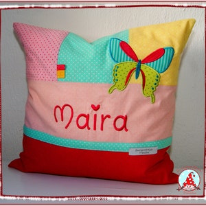 Könnte beinhalten: Ein rotes, rosa, gelbes und grünes Patchwork-Kissen mit einer Schmetterlingsapplikation und dem Namen "Maira" in rot gestickt. Das Kissen hat ein weißes Herz über dem Namen.
