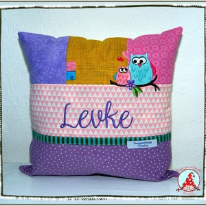 Könnte beinhalten: Ein lila, pink und gelbes Kissen mit Patchwork-Design. Das Kissen zeigt zwei gestickte Eulen und den Namen "Levke".