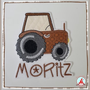 Könnte beinhalten: Ein gestickter Stoffaufnäher mit einem braunen Traktor, einer cremefarbenen Kabine und dunkelgrauen, strukturierten Rädern. Unter dem Traktor ist der Name MORITZ in braunem Garn gestickt, mit einem kleinen Stern im Buchstaben O.