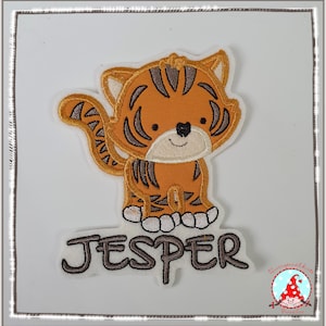 Könnte beinhalten: Eine gestickte Tiger-Applikation in Braun und Orange auf weißem Hintergrund. Der Tiger steht auf seinen Hinterbeinen, der Schwanz ist um seinen Körper gewickelt. Der Name "Jesper" ist unter dem Tiger in braunem Garn gestickt.