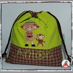 Könnte beinhalten: Grüne Kordelzugtasche mit braunem kariertem Boden. Die Tasche hat ein gesticktes Design mit einem Mädchen, das einen grünen Hut trägt, und einem Affen. Der Name Anton ist auf die Tasche gestickt.