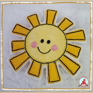 Könnte beinhalten: Ein gelber, sonnenförmiger Stoffaufnäher mit einem lächelnden Gesicht, schwarzen gestickten Augen, einem geschwungenen Mund und zwei rosa Wangen. Die Sonne hat zwölf strahlenförmige, rechteckige Zacken, die mit schwarzem Garn auf weißem Grund umrandet sind.