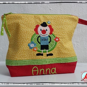 May include: Yellow fabric cosmetic bag with a clown applique. The bag has a green zipper and a red base with the name "Anna" embroidered in green. The clown has red hair, a black hat, and the word "b&auml;h!" on its shirt.