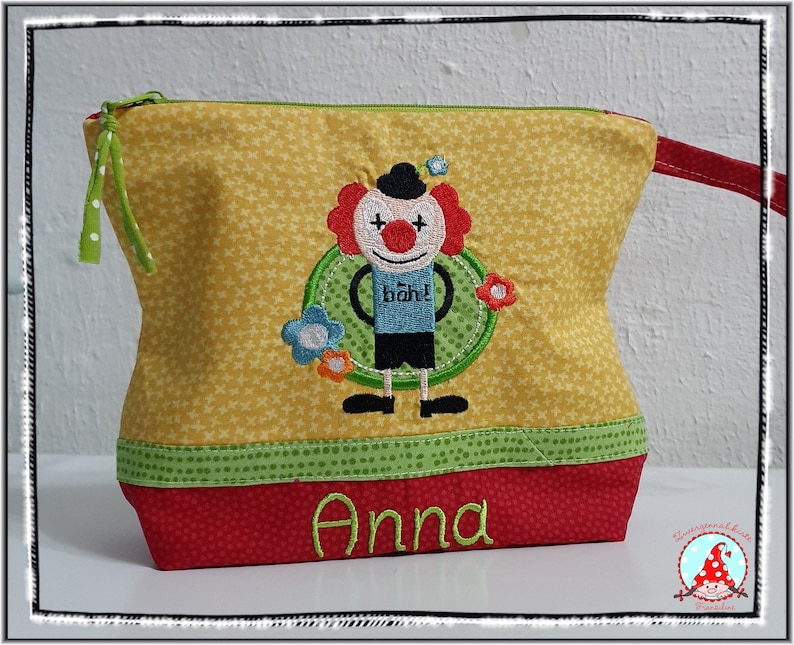 May include: Yellow fabric cosmetic bag with a clown applique. The bag has a green zipper and a red base with the name "Anna" embroidered in green. The clown has red hair, a black hat, and the word "b&auml;h!" on its shirt.