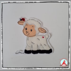 Könnte beinhalten: Ein weißes Filzlamm mit einem roten Marienkäfer auf dem Kopf. Das Lamm hat schwarze Filzfüße und ein rotes Herz auf dem Rücken.