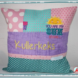 Könnte beinhalten: Ein Patchwork-Kissen mit verschiedenen Mustern und Farben. Der obere Bereich zeigt rosa Punkte und Herzen, mit dem gestickten Text "You are my sunshine" und "Kullerkecks". Der untere Bereich ist türkis mit einem floralen Design.