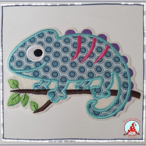 Könnte beinhalten: Ein gestickter Chamäleon in Blau und Grün mit einem rosa Streifen und lila Flecken, der auf einem braunen Ast mit grünen Blättern sitzt. Das Chamäleon hat ein weißes Auge mit einer schwarzen Pupille.