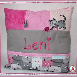 Könnte beinhalten: Rosa-graues Kissen mit einer Katze, die mit Garn spielt, und dem Namen "Leni" darauf gestickt. Das Kissen hat ein Patchwork-Design mit rosa Punkten, rosa Streifen und einem grauen Stoff mit Katzenmuster.