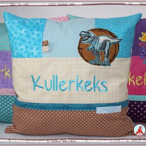 Könnte beinhalten: Ein hellblaues, beiges und braunes Kissen mit einem Dinosaurier-Motiv und dem Namen "Kullerkeks" bestickt.