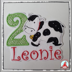 Könnte beinhalten: Grüne und weiße gestickte Zahl Zwei mit einer schwarzen und weißen Kuh-Applikation und dem Namen Leonie in Rot gestickt.