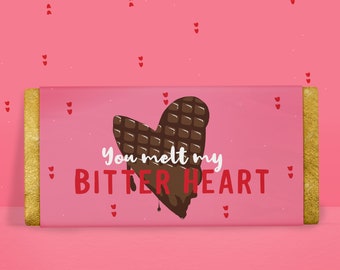 May include: A pink chocolate bar wrapper with gold foil ends. The wrapper features a dark chocolate heart design with the text "You melt my BITTER HEART" in white and red. Small red hearts are scattered on the pink background.