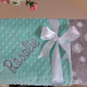 Pode incluir: Um cobertor para bebé de cor verde menta com uma fita de cetim branca amarrada em um laço. O cobertor é personalizado com o nome "Rosalie" em bordado cinza. O outro lado do cobertor é cinza com um padrão de dentes-de-leão brancos.