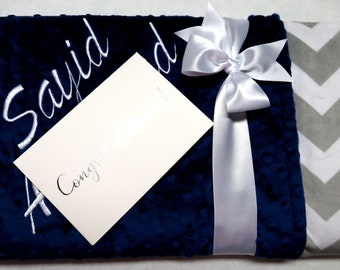 May include: A navy blue and gray chevron baby blanket with the name "Sayid" embroidered on it. The blanket is tied with a white satin ribbon and includes a white card with the word "Congrats" written on it.