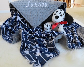 Puede incluir: Una manta para bebé gris y azul con un patrón de flechas blancas. La manta está personalizada con el nombre "Jaxson" y presenta un tejido minky gris con puntos en un lado. Un juguete de peluche de cachorro dálmata está acostado sobre la manta.
