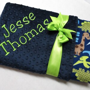 May include: A navy blue baby blanket with a green ribbon and the name "Jesse Thomas" embroidered in green. The other side of the blanket features a blue and green dinosaur print.