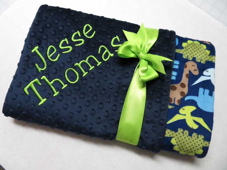May include: A navy blue baby blanket with a green ribbon and the name "Jesse Thomas" embroidered in green. The other side of the blanket features a blue and green dinosaur print.