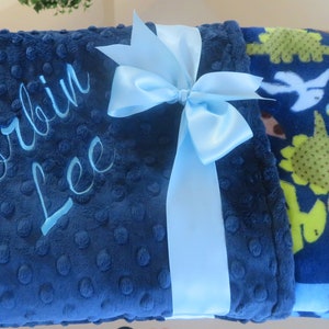 May include: A blue baby blanket with a light blue satin ribbon tied in a bow. The blanket is personalised with the name "Korbin Lee" embroidered in light blue thread. One side of the blanket features a blue background with a pattern of green and yellow dinosaurs.