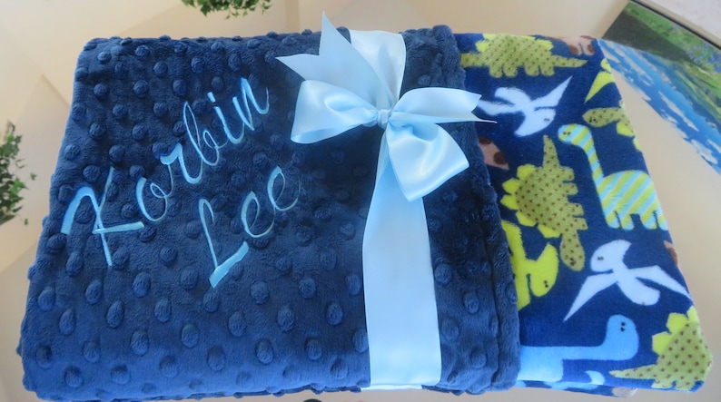 May include: A blue baby blanket with a light blue satin ribbon tied in a bow. The blanket is personalised with the name "Korbin Lee" embroidered in light blue thread. One side of the blanket features a blue background with a pattern of green and yellow dinosaurs.
