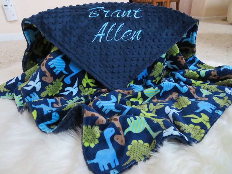 May include: A blue and green baby blanket with a dinosaur print on one side and a navy blue minky dot fabric on the other side. The blanket is personalised with the name "Brant Allen" embroidered on the navy blue side.