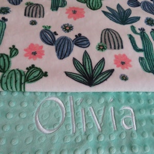 May include: A white and green baby blanket with a cactus and flower pattern. The blanket has a minky side and a soft, textured side. The name "Olivia" is embroidered on the textured side.