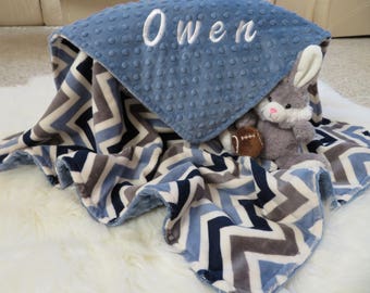 May include: A blue and gray chevron patterned baby blanket with a blue minky dot fabric on the top. The blanket is personalized with the name "Owen" in white embroidery. A gray plush bunny with a football is lying on the blanket.