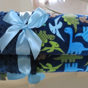May include: A blue and green baby blanket with a dinosaur print. The blanket is rolled up and tied with a light blue ribbon.
