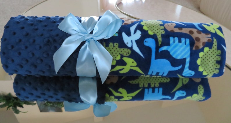 May include: A blue and green baby blanket with a dinosaur print. The blanket is rolled up and tied with a light blue ribbon.