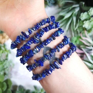 May include: Several blue lapis lazuli chip bead bracelets are stacked on a wrist. The bracelets are made of small, irregular pieces of the deep blue stone. The bracelets are strung together, creating a layered look. The background features green plants.