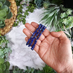 May include: Three strands of blue lapis lazuli gemstone beads are held in a hand. The beads are small, irregular chips of deep blue color. The background includes green plants and a woven basket.
