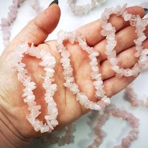 May include: A hand holding a strand of pink rose quartz chip beads. The beads are strung together to form a bracelet or necklace. The beads are irregular in shape and size.