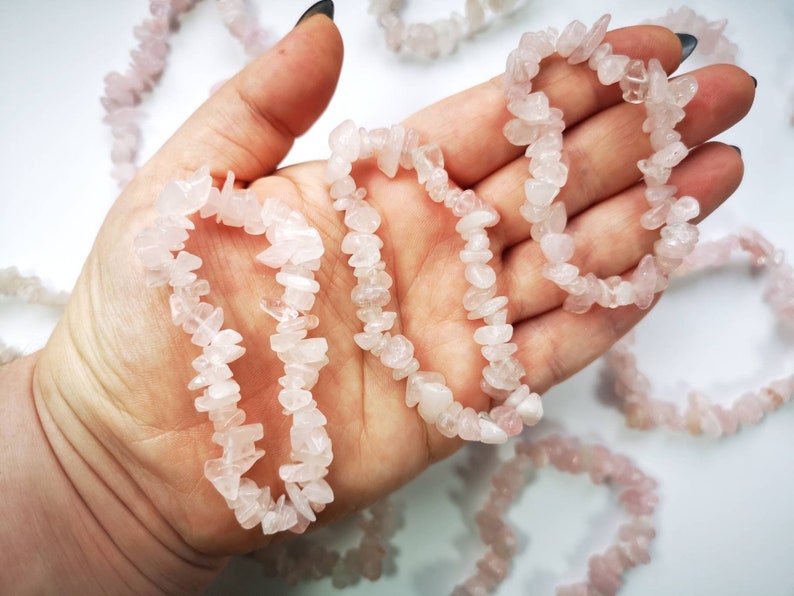 May include: A hand holding a strand of pink rose quartz chip beads. The beads are strung together to form a bracelet or necklace. The beads are irregular in shape and size.
