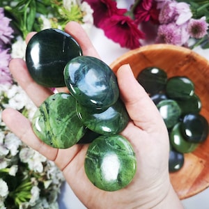 May include: A selection of polished, oval green stones, some with darker green veining, are held in a hand and displayed in a wooden bowl. The stones have a smooth, glossy surface. The background includes white and purple flowers.