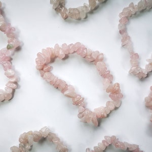 May include: A collection of six pink rose quartz chip bracelets. Each bracelet is made of small, irregular shaped chips of rose quartz, strung together on a clear elastic cord.