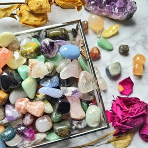 May include: A collection of polished gemstones and crystals in various colours, such as amethyst and rose quartz, are presented in a clear glass tray. Additional stones and a rose are scattered around the tray on a white marble surface. The stones vary in size and shape.