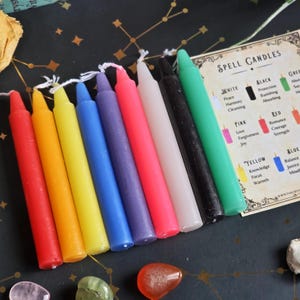 May include: A selection of multi-coloured spell candles, including red, orange, yellow, blue, purple, pink, white, black, and green. Each candle has a wick. A card with text about spell candles is visible.