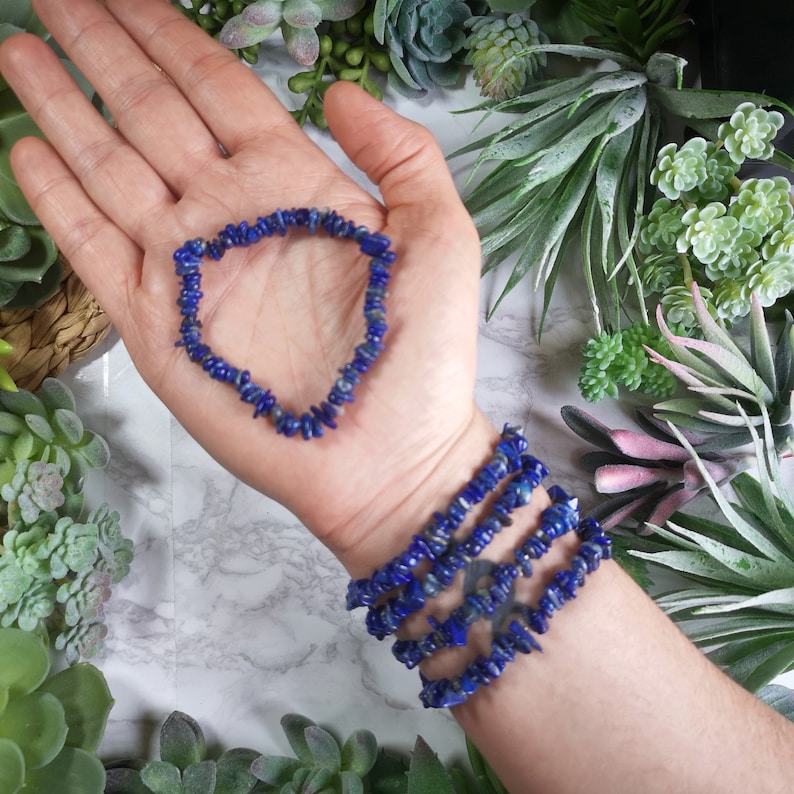 May include: A collection of blue lapis lazuli bracelets. One bracelet is held in an open palm, while several others are stacked on a wrist. The bracelets are made of small, irregularly shaped beads. The background features green and pink plants.